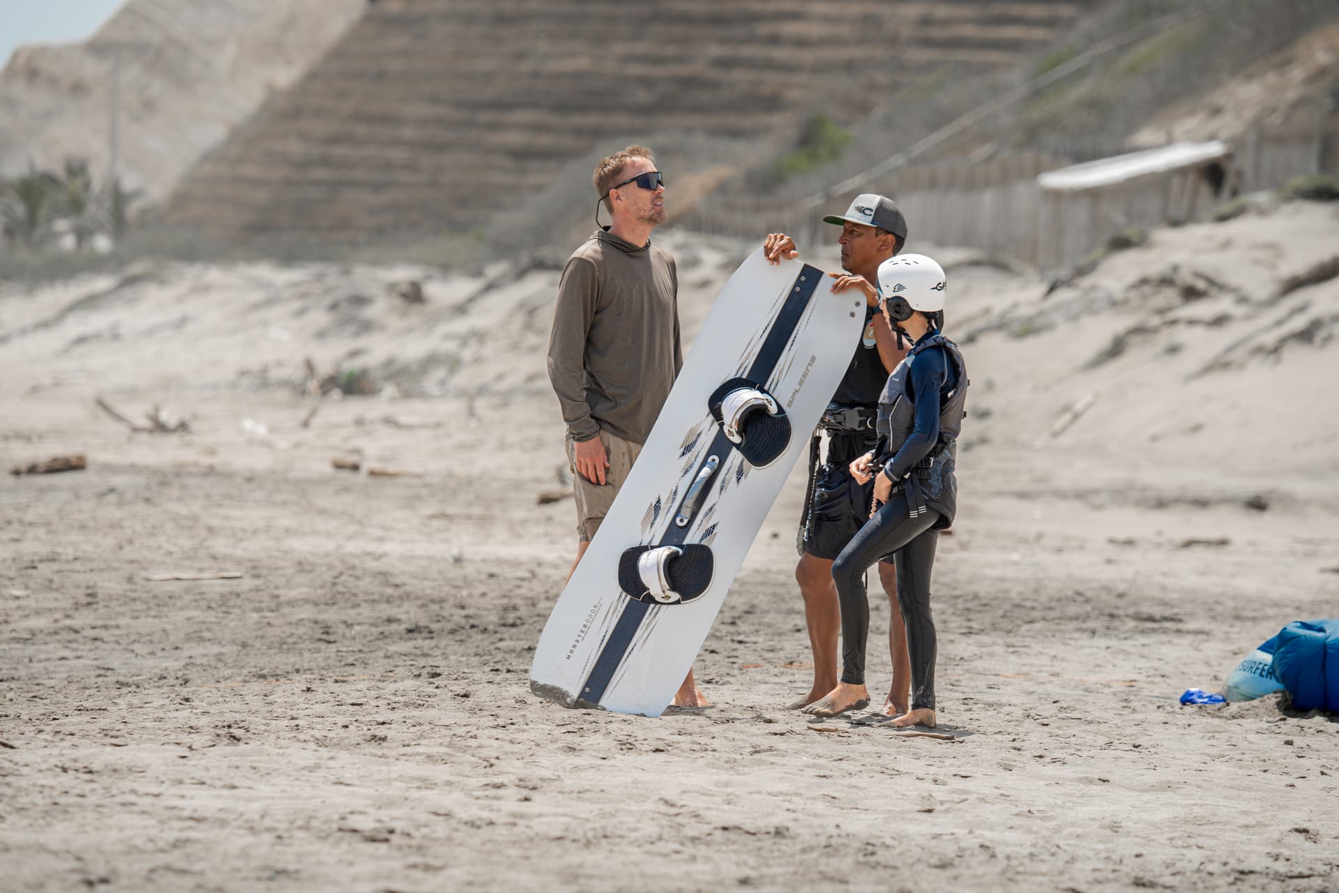 A kitesurf instructor briefing a student next to a board planted in the sand